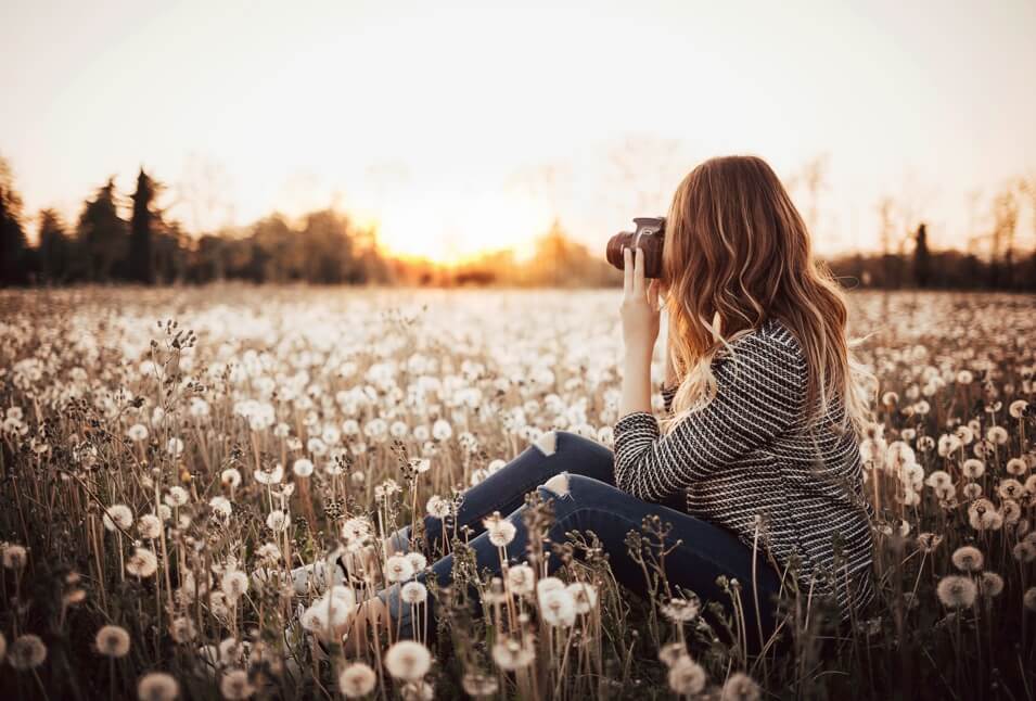A woman sits on the ground surrounded by dandelions and takes picture of nature