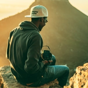Dark skin man sits on the rock with a camera, surrounded by mountains